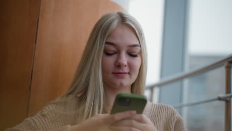 Close-up of young lady operating iPhone with green case while seated at wooden table. Her hands are focused on screen, showing attention to detail in a relaxed environment