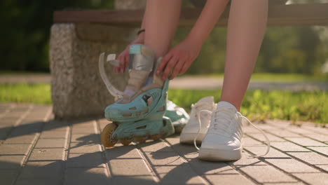 lower view of person putting on roller skate on right leg with other roller skate slightly blurred in background, sneakers close by on pavement, warm sunlight casting shadows, greenery in background