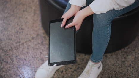 hands holding a tablet, gently tilting it with soft reflections on the screen, casual outfit with denim jeans and white sneakers emphasizes modern lifestyle in subtle lighting and reflective flooring