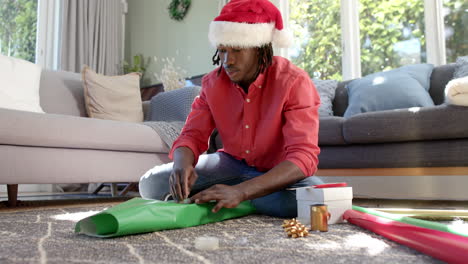 happy african american man in christmas hat wrapping gift on floor at home, slow motion