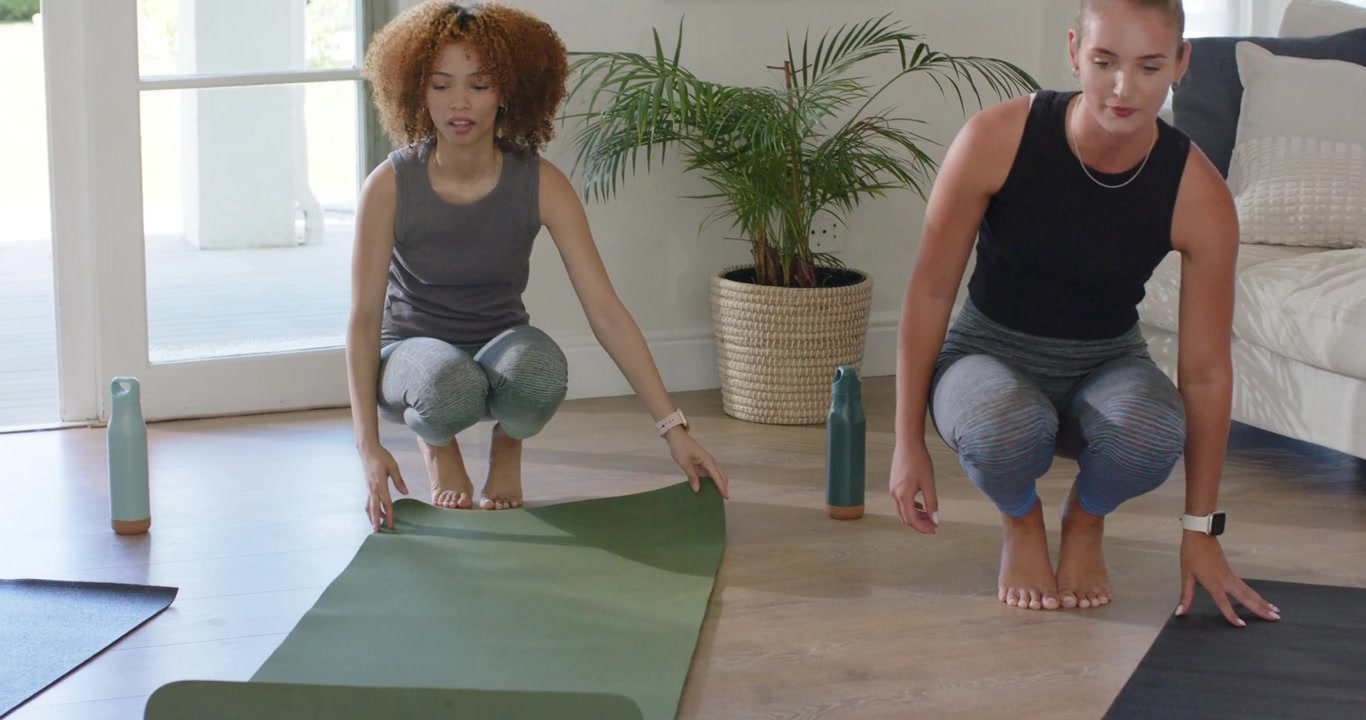 Partner di yoga donne si preparano per esercizi a terra, inginocchiate mentre srotolano i tappetini a casa e posizionano le bottiglie.