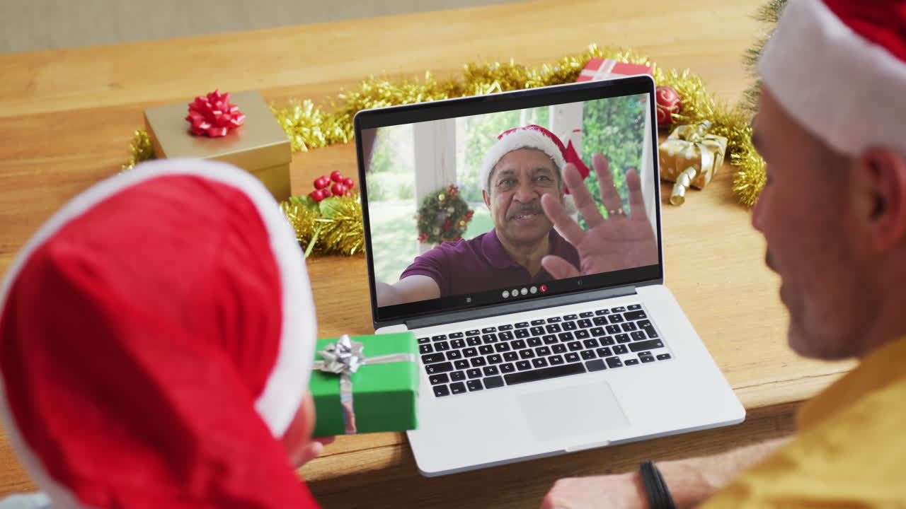 padre caucásico con hijo con sombreros de santa usando una computadora portátil para una videollamada de navidad, con el hombre en la pantalla
