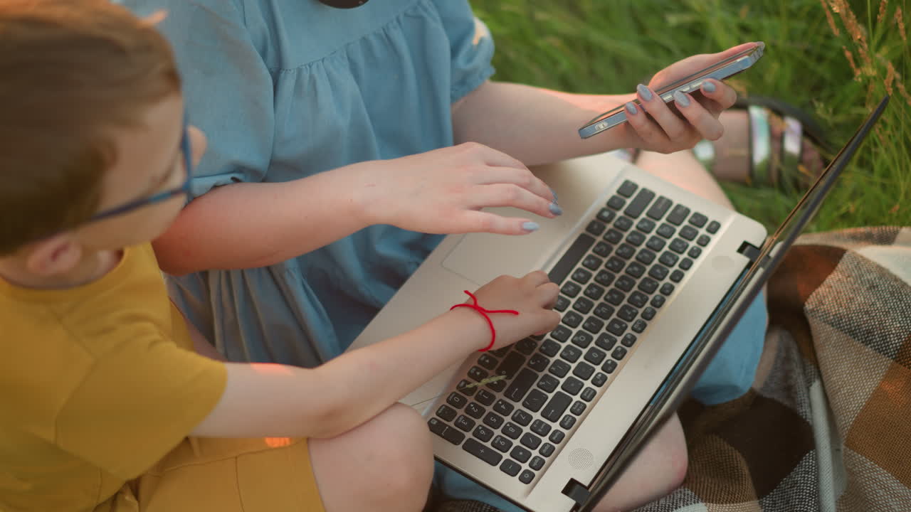 un primer plano de una mujer sentada en la hierba con una computadora portátil en el regazo, usando simultáneamente su teléfono. un niño joven, vestido de amarillo con una banda roja en la muñeca, extiende la mano para tocar el teclado
