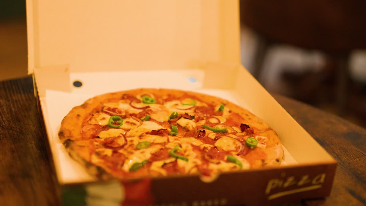 Hand opens pizza box revealing hot pizza under warm indoor lighting, shallow depth of field