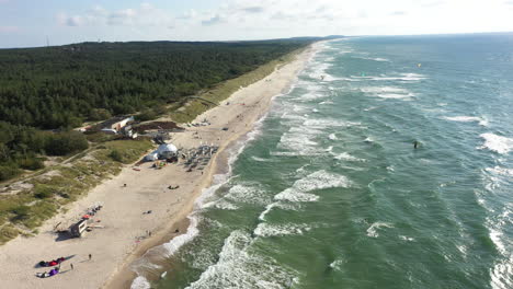 AERIAL:-Rotating-Shot-of-Surfers-Riding-Waves-with-Kites-on-a-Sunny-Day-with-Forest-and-Bright-Sky-in-Background