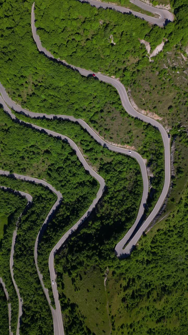 Drammatica ripresa aerea dall'alto di una strada di montagna asfaltata e tortuosa con numerosi tornanti, che si snoda attraverso un paesaggio verde vibrante e densamente boscoso. Le auto sono visibili mentre affrontano le impegnative curve.