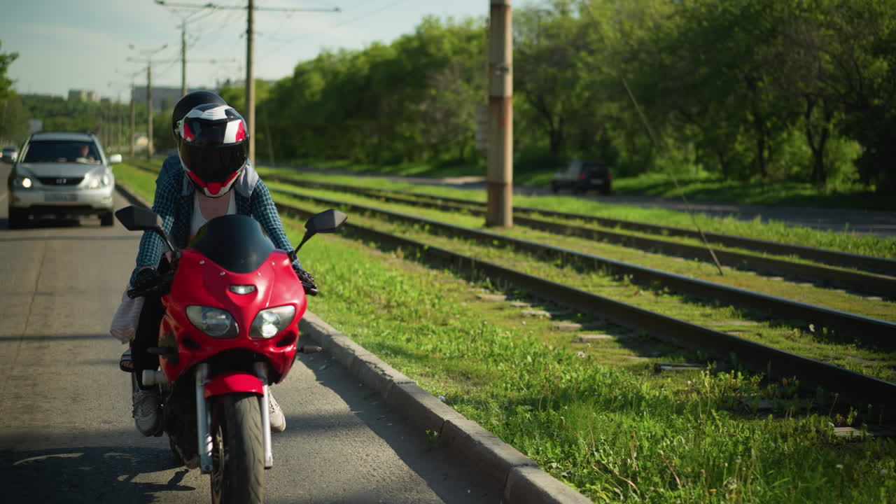 dos amigos montan una bicicleta eléctrica roja lentamente con cascos, cerca de una vía ferroviaria, un coche sigue detrás de ellos, y árboles verdes exuberantes se alinean en el lado opuesto de la carretera