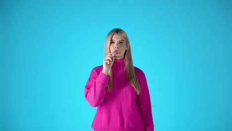 Caucasian-young-blonde-woman-with-pink-sweater-eats-a-cookie-staring-at-camera-in-blue-chroma-background,-torso-view