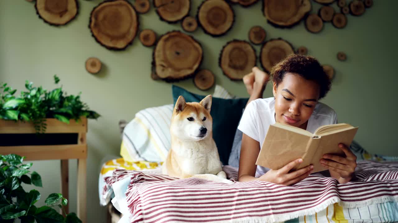sonriente estudiante afroamericana chica encantadora está leyendo un libro en la cama en casa mientras su perro mascota está acostado cerca de ella. pasatiempo, ocio, animales y concepto de interior.
