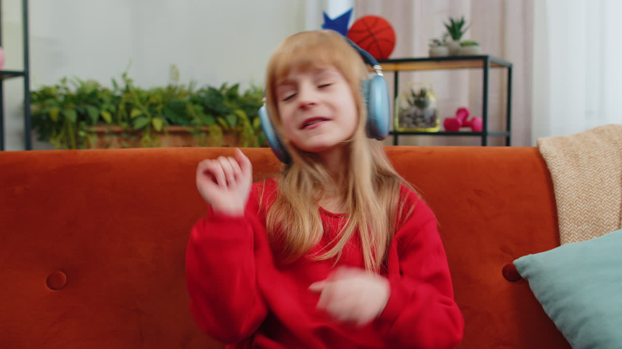 Happy teen girl in wireless headphones relaxing sitting on sofa at home listening favorite music
