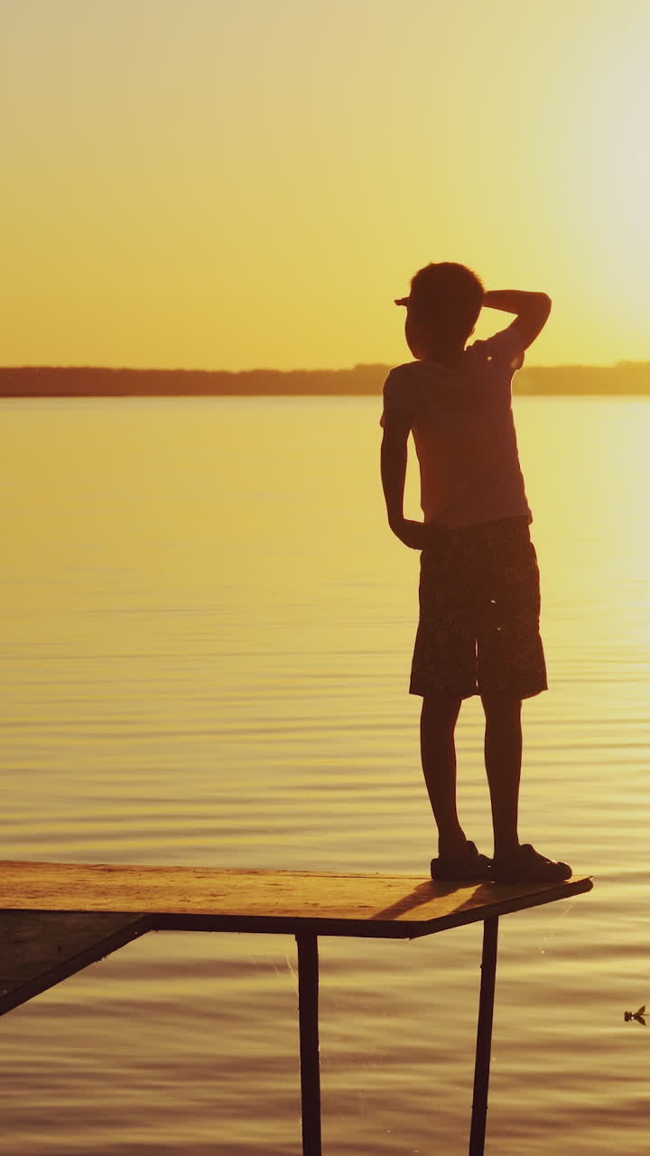 A little kid is standing on the masonry near the bench. The guy is looking around the area at sunset near the lake in the summer. Vertical video