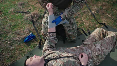 a confident male military man in a camouflage green uniform ties a fabric bandage to the unconscious hand of a male soldier in a combat area outside the city