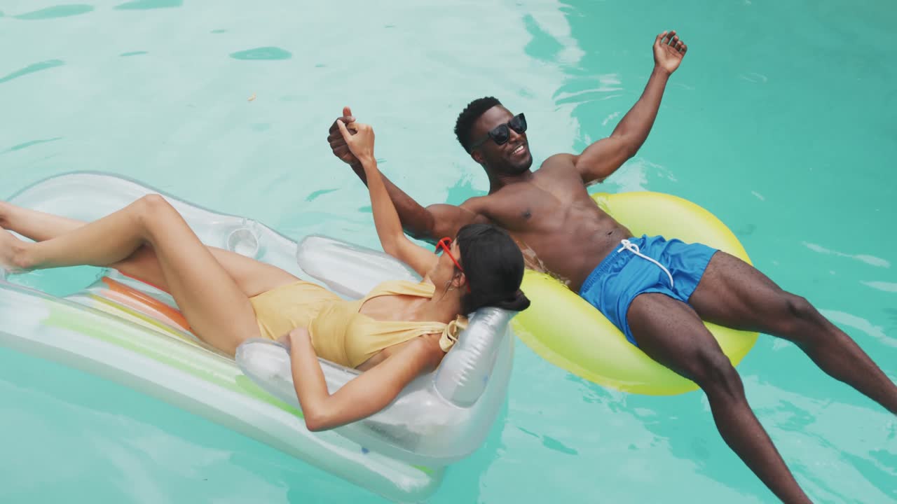 Happy diverse couple wearing swimming suits with inflatables at swimming pool in garden