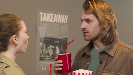 happy couple holding popcorn box and drinking soft drink while talking together at the cinema