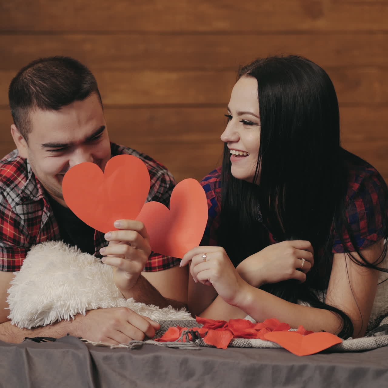 Happy couple lying on bed and showing different grimaces closing their faces by red paper hearts. Man and woman hold paper hearts on faces, look at camera and do passionate kiss.
