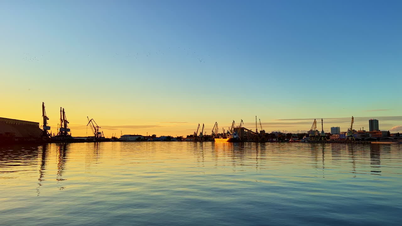 Sunset over the harbor reflecting calm waters while cranes and boats create a serene maritime scene in the late evening glow