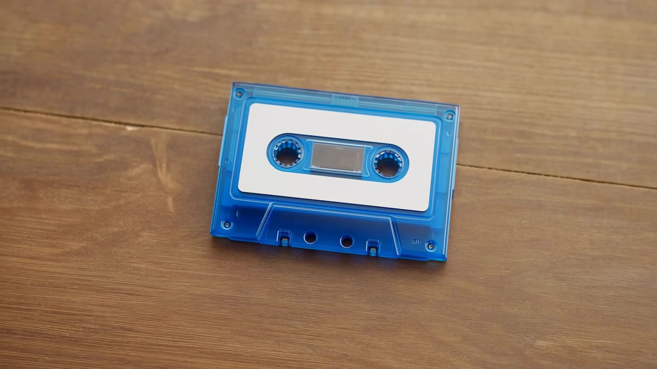 Composizione dall'alto che mostra una cassetta audio blu trasparente appoggiata vicino al centro superiore di un tavolo di legno marrone caldo. L'involucro di plastica trasparente rivela