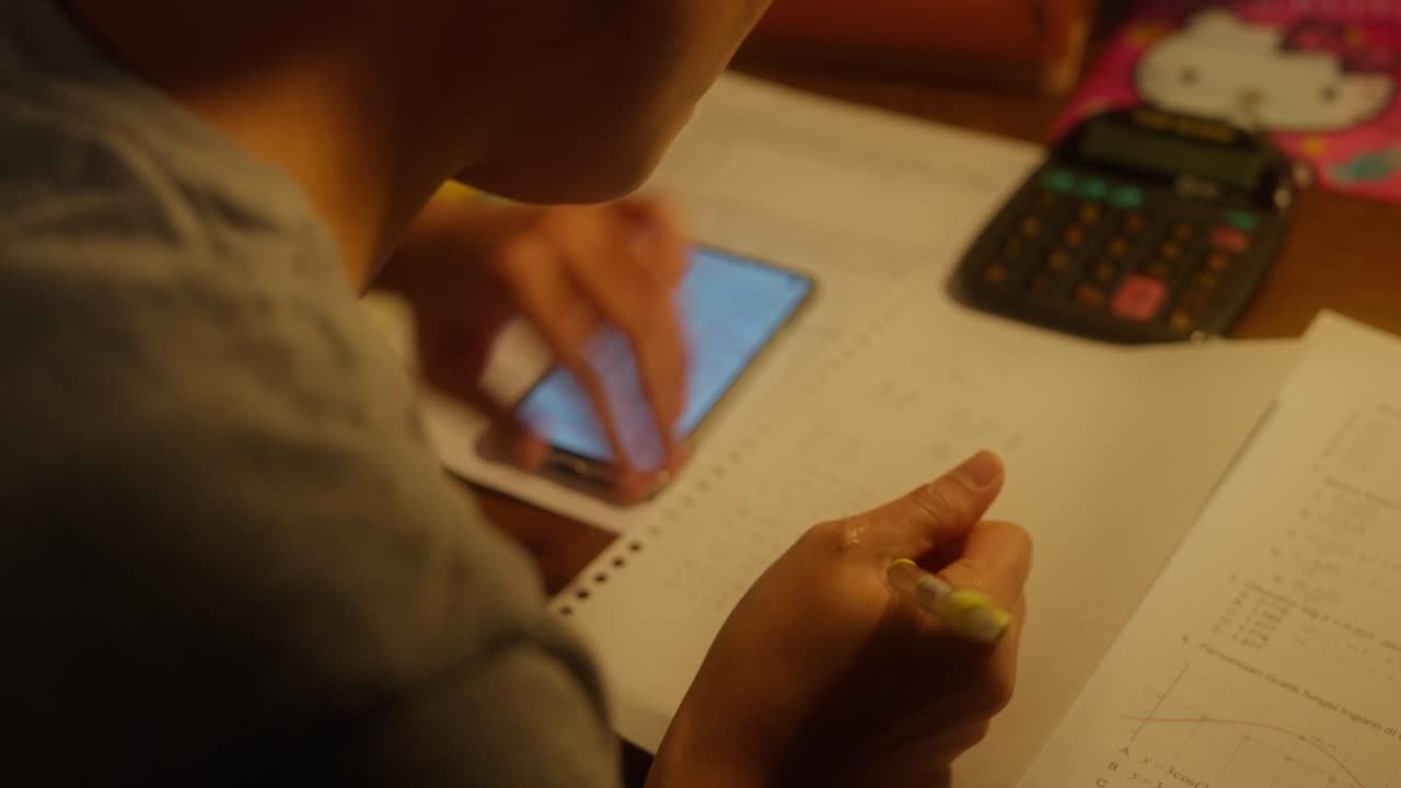 primer plano de una niña estudiando escribiendo tareas de matemáticas en un escritorio en una habitación con una cálida luz de tungsteno usando un teléfono