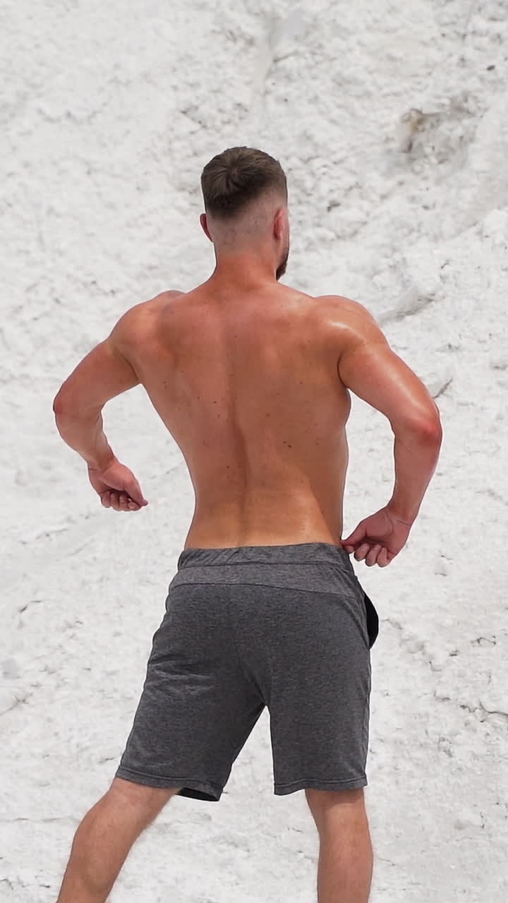 Backside view of a bodybuilder outdoors. Strong athlete without shirt shows his muscular body on white mountain background. Slow motion. Vertical video