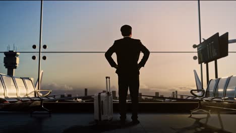 full body back view of asian businessman with rolling suitcase in boarding lounge at the airport, businessman with arms akimbo looking around waits for flight, airplane takes off outside the window