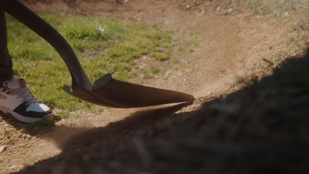 Man uses shovel in outdoor field under bright daylight, preparing soil in a rural garden setting