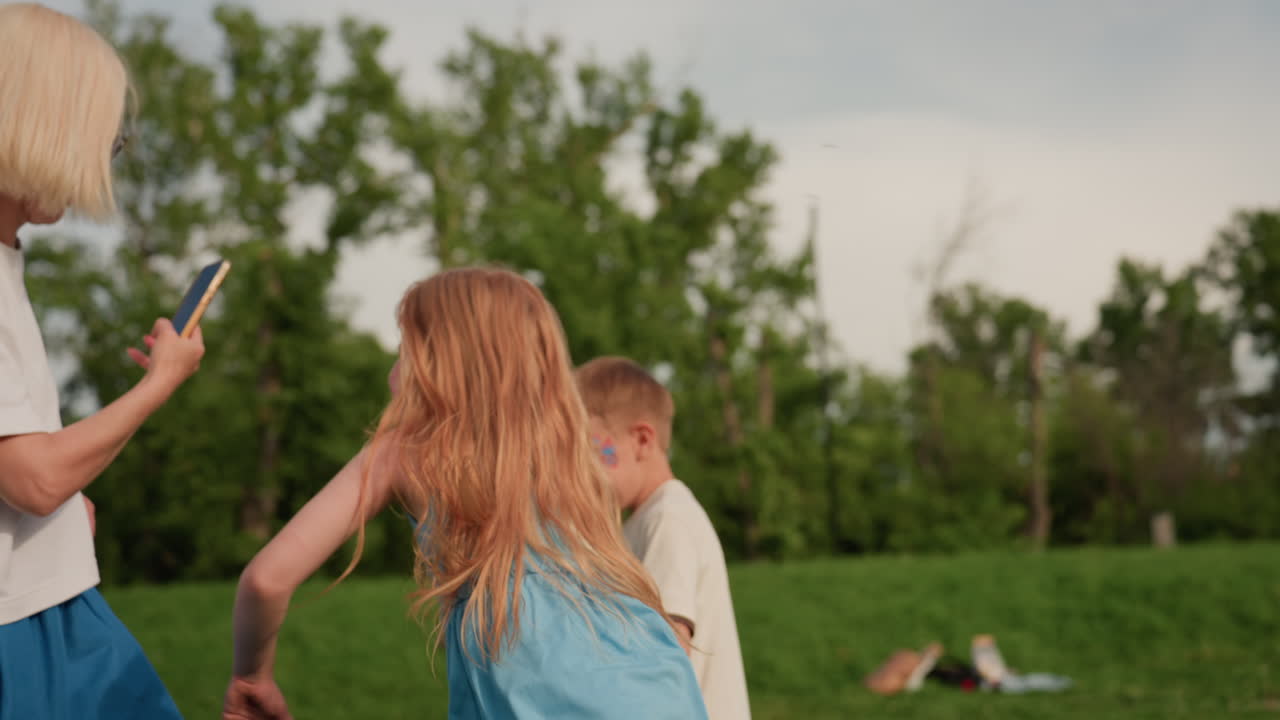 holiday kids running toward parents videoing them on phone, smiling, holding hands, wind in hair, playful energy in green park grass on summer afternoon, carefree childhood motion and joy