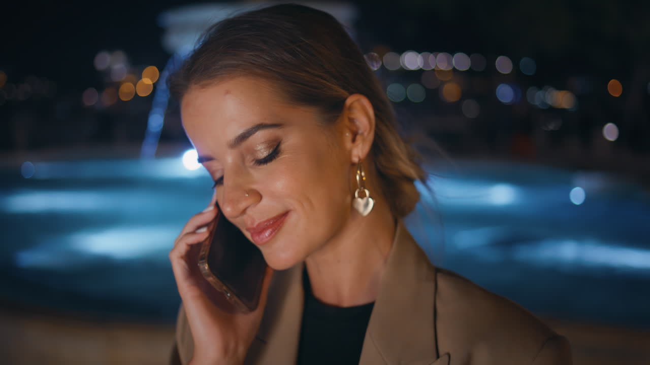 en primer plano, una mujer hermosa hablando por teléfono celular por la noche con una fuente de agua iluminada.