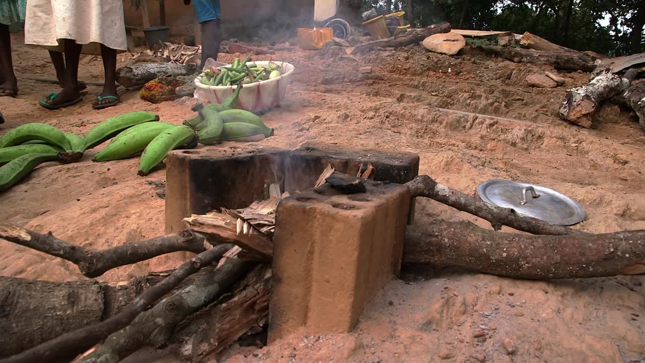 Area di cottura tradizionale all'aperto nel Ghana rurale con una stufa a legna, fumo e legna da ardere, e banane plantano verdi fresche pronte per la preparazione del cibo in un ambiente di villaggio.