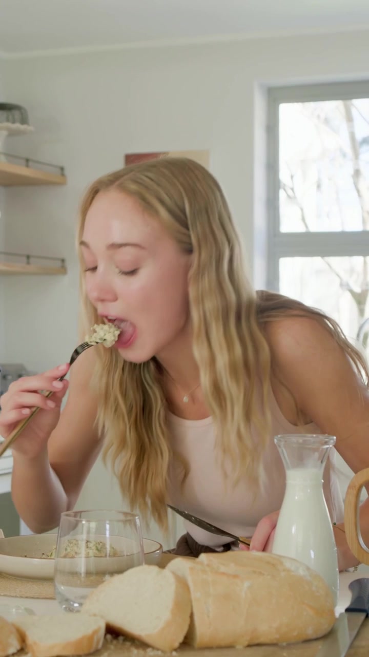 Video vertical: Empezando a cortar, mujer con top color canela probando comida en la cocina con pan, jarra de leche