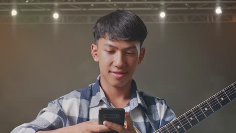 close up of asian teenager with guitar using smartphone while standing on stage