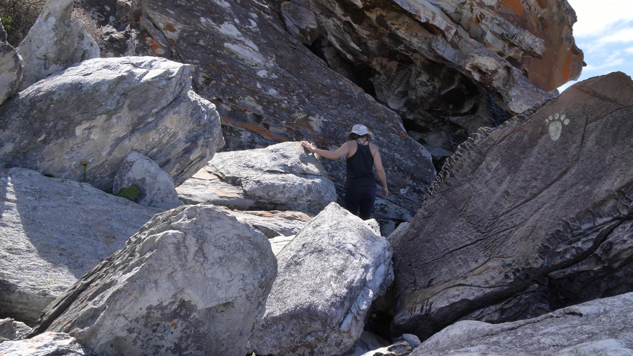 Two people navigate a challenging, sun-drenched, rocky landscape, discovering a unique painted paw print on a large boulder at the Garden Route National Park, Tsitsikamma, Western Cape.