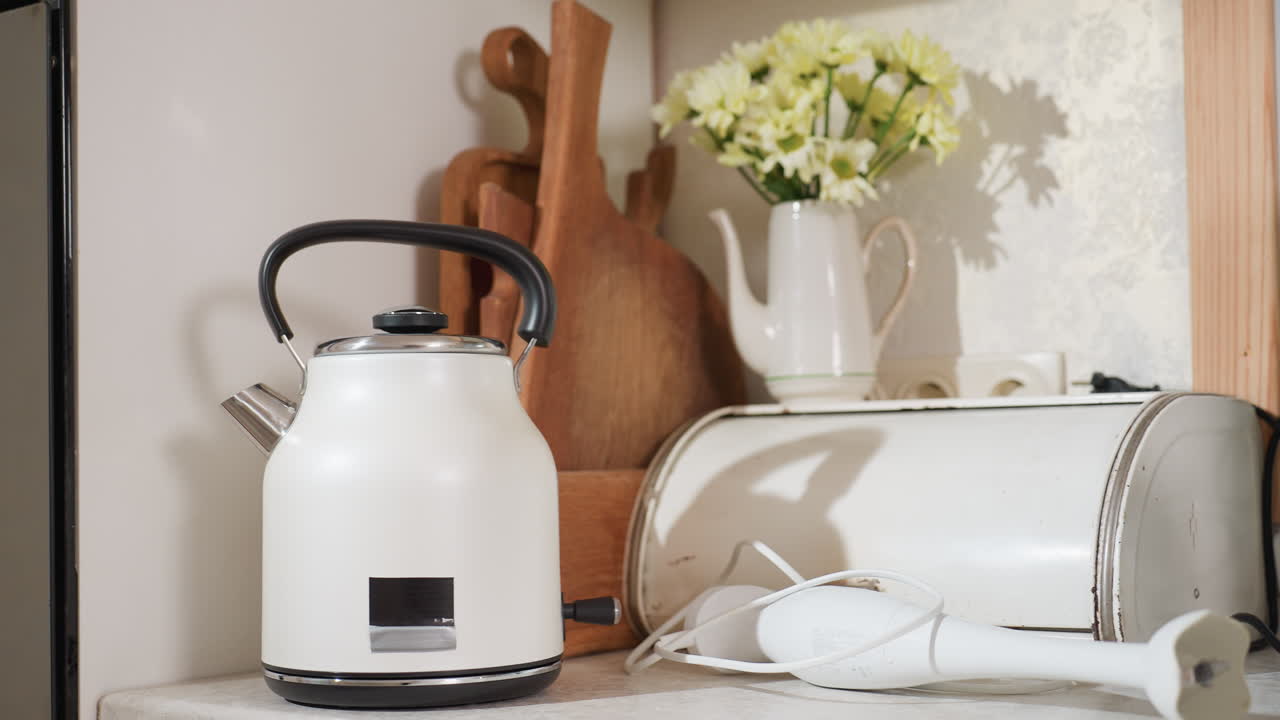 White electric kettle on kitchen counter beside wooden chopping boards, stainless appliances, electric hand blender, and ceramic jug with flowers, all arranged in bright homely kitchen