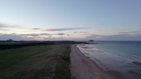 Imágenes-De-Drones-Que-Vuelan-Lentamente-Hacia-Atrás-Sobre-Una-Larga-Playa-De-Arena-Y-Dunas-De-Arena-Durante-Una-Puesta-De-Sol-Rosa-Mientras-La-Marea-Golpea-Suavemente-La-Orilla-Para-Revelar-Que-La-Gente-Camina-Por-La-Playa