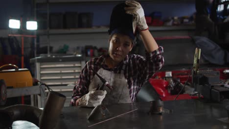 Portrait-of-female-mechanic-removing-welding-helmet-while-sitting-at-a-car-service-station