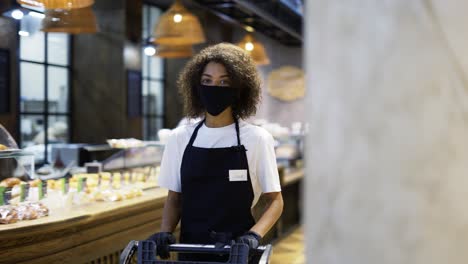 african american female worker pushing trolley in supermarket, wearing protection mask