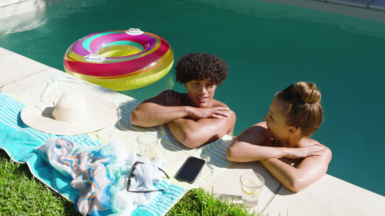 Relaxing by pool, diverse couple enjoying sunny day with drinks and inflatable ring
