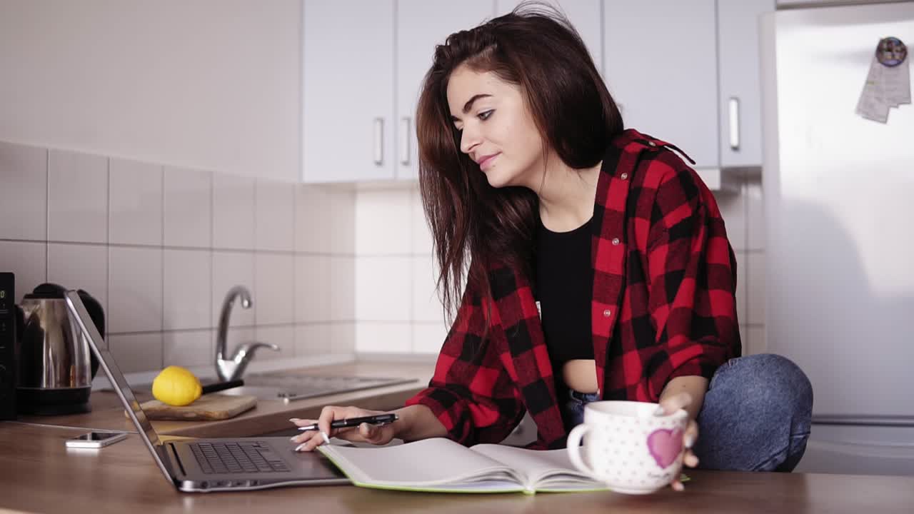 Attractive girl in her 20's looks forsomething on the internet using her laptop and then writes something down in her notebook while sitting in a cozy kitchen of her apartment