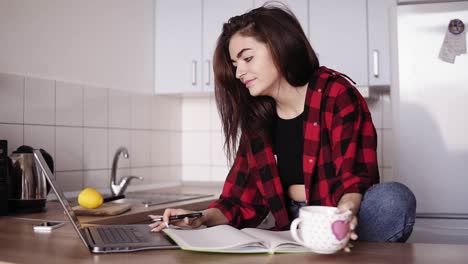 chica atractiva de unos 20 años busca algo en internet usando su portátil y luego escribe algo en su cuaderno mientras está sentada en una cocina acogedora de su apartamento