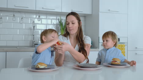 madre de familia joven y dos hijos sentados en la cocina en la mesa blanca comiendo hamburguesas juntos y divirtiéndose. los niños ríen en la mesa de la cena