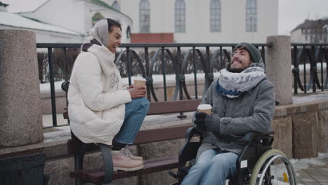 muslim woman and her disabled friend in wheelchair drinking takeaway coffe on a bench in city in winter 2