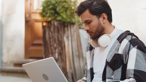 indian young man freelancer sitting on street using laptop typing working online distant job outdoor