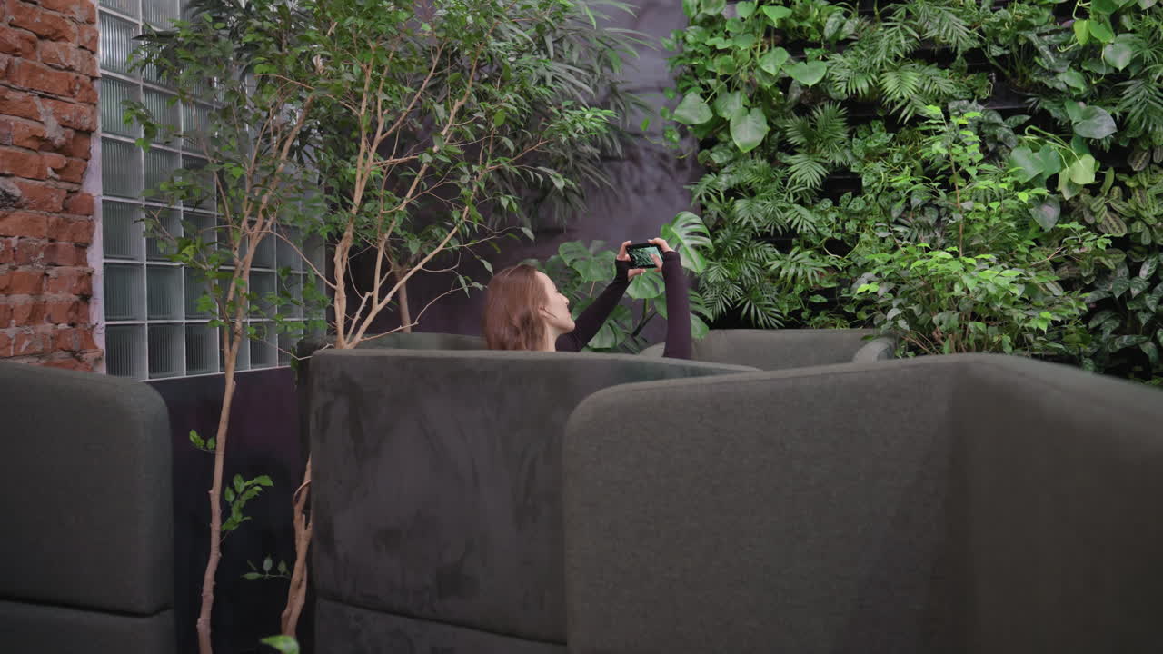 Lady positioned between soft green cushions lifts phone to take picture in plant-filled lounge with leafy vertical wall and indoor trees adding serene, natural atmosphere to modern cozy space