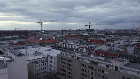Forwards-fly-above-buildings-in-urban-neighbourhood.-Group-of-tower-cranes-against-overcast-sky.-Berlin,-Germany