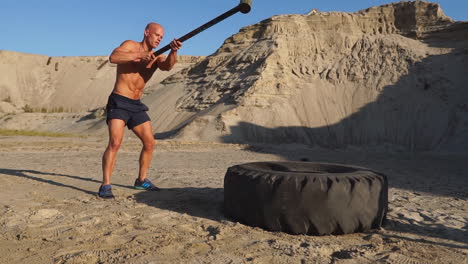 atleta musculoso hombre fuerte golpea un martillo en una rueda enorme en las montañas arenosas en cámara lenta al atardecer. el polvo de las ruedas se eleva.