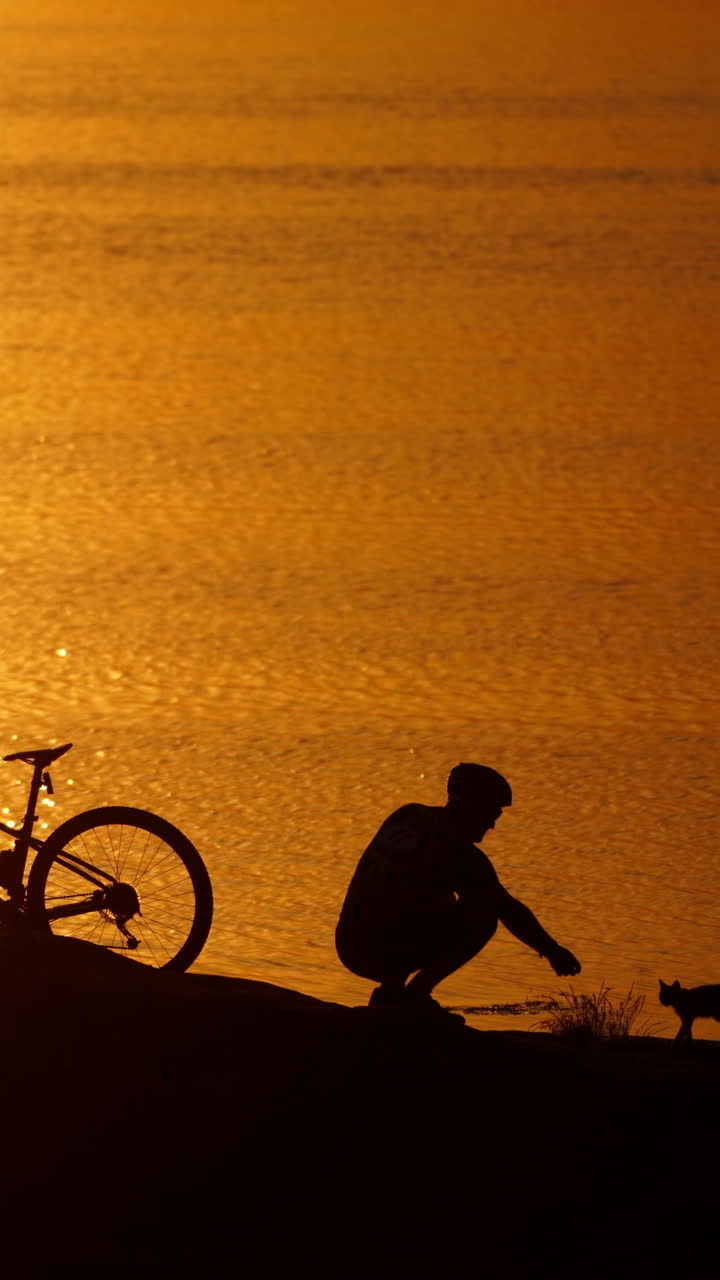 Man with bicycle resting on beach. Silhouette of man resting after cycling trip near river Vertical video