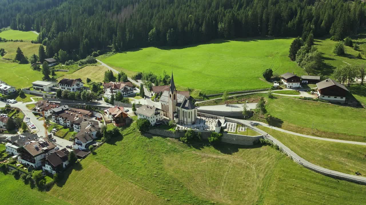 An aerial view of a village featuring a historic church with a tall spire, surrounded by residential houses and lush green alpine fields. Dense forest borders the village.