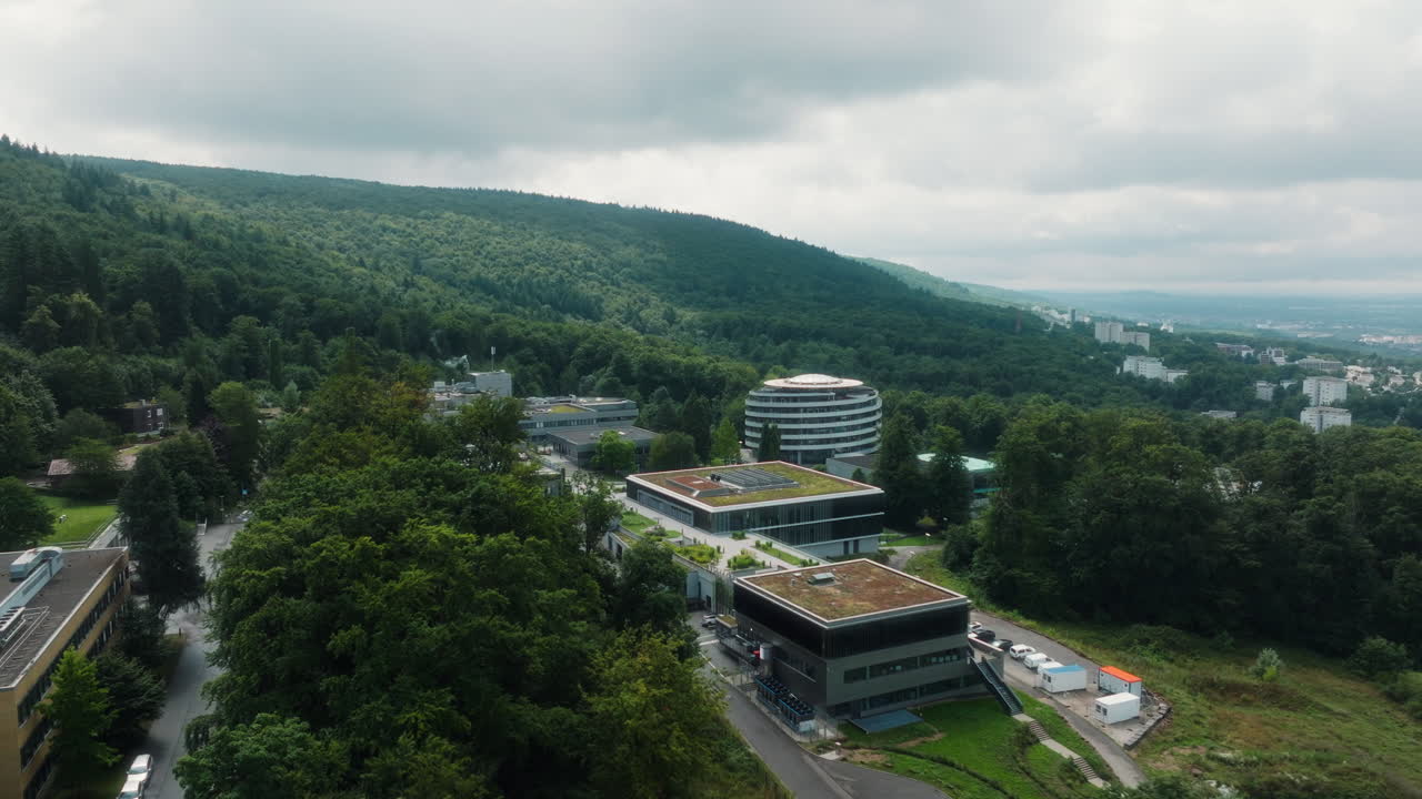 Drone flying forward over forested hillside toward DKFZ buildings with cloudy sky