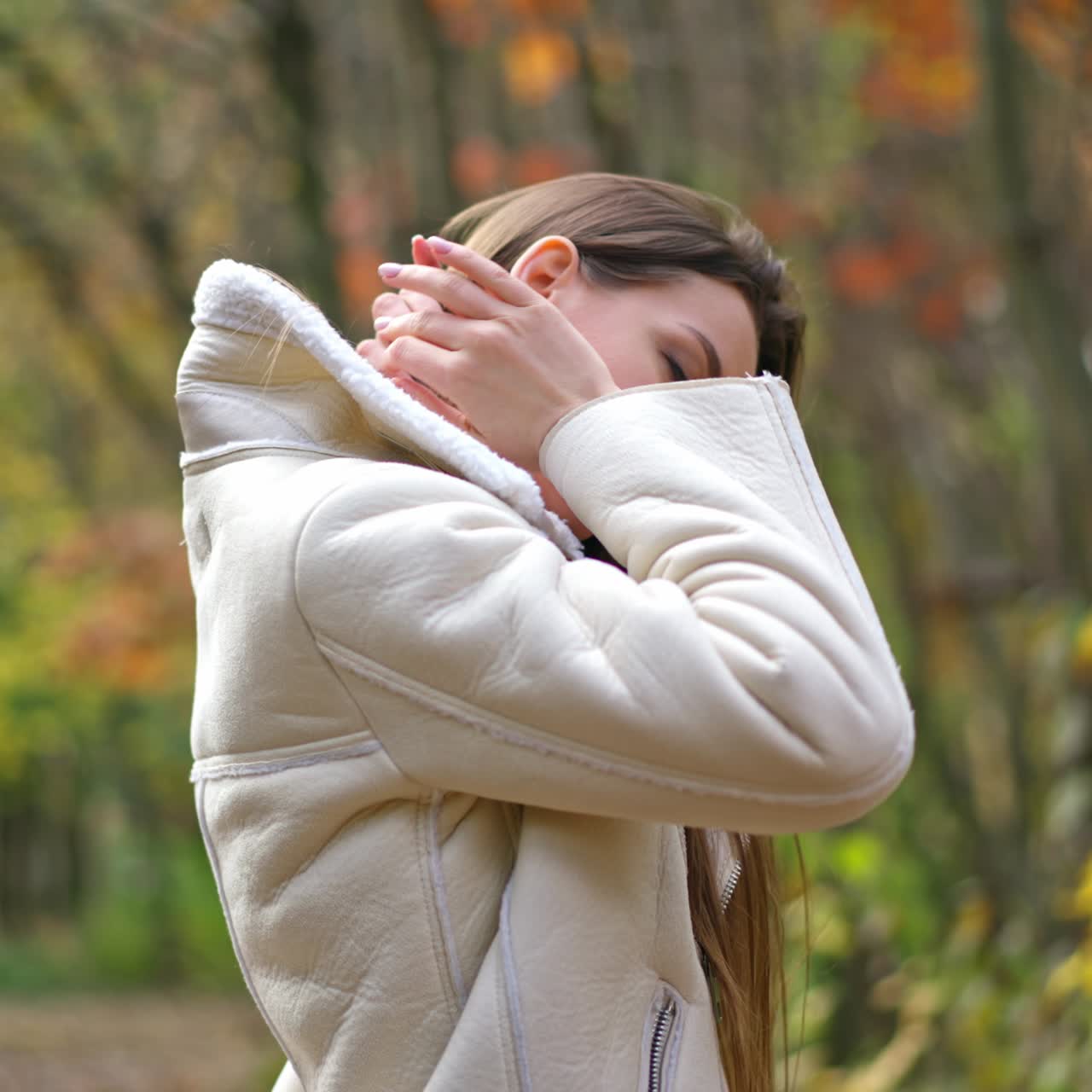 Relaxed calm woman wearing warm jacket touching long hair. Smiling lady at the walk along the autumn colorful park