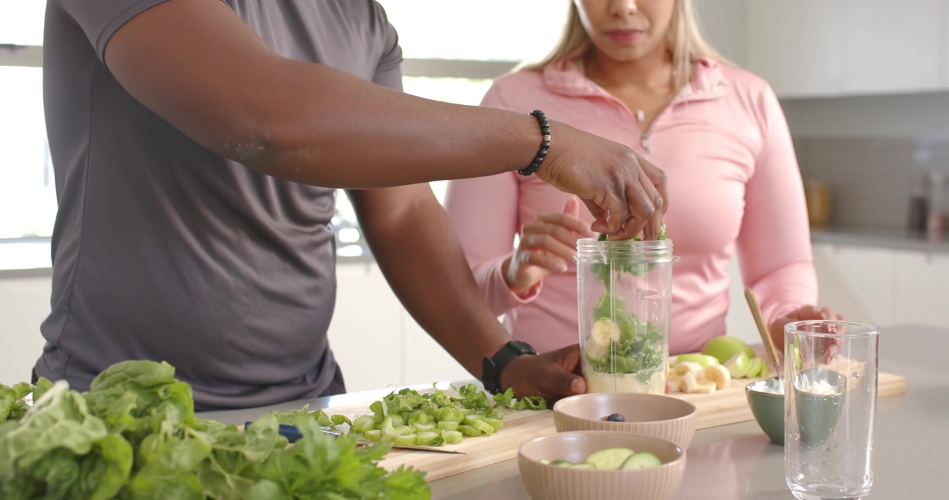 Casal afro-americano picando verduras na cozinha de casa, adicionando ervas no copo do liquidificador para fazer uma bebida.