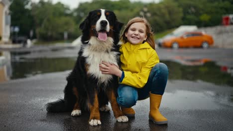 Porträt-Eines-Glücklichen-Blonden-Mädchens-In-Einer-Gelben-Jacke,-Das-Neben-Ihrem-Großen-Schwarz-weißen-Rassehund-Und-Im-Park-Nach-Dem-Regen-Sitzt